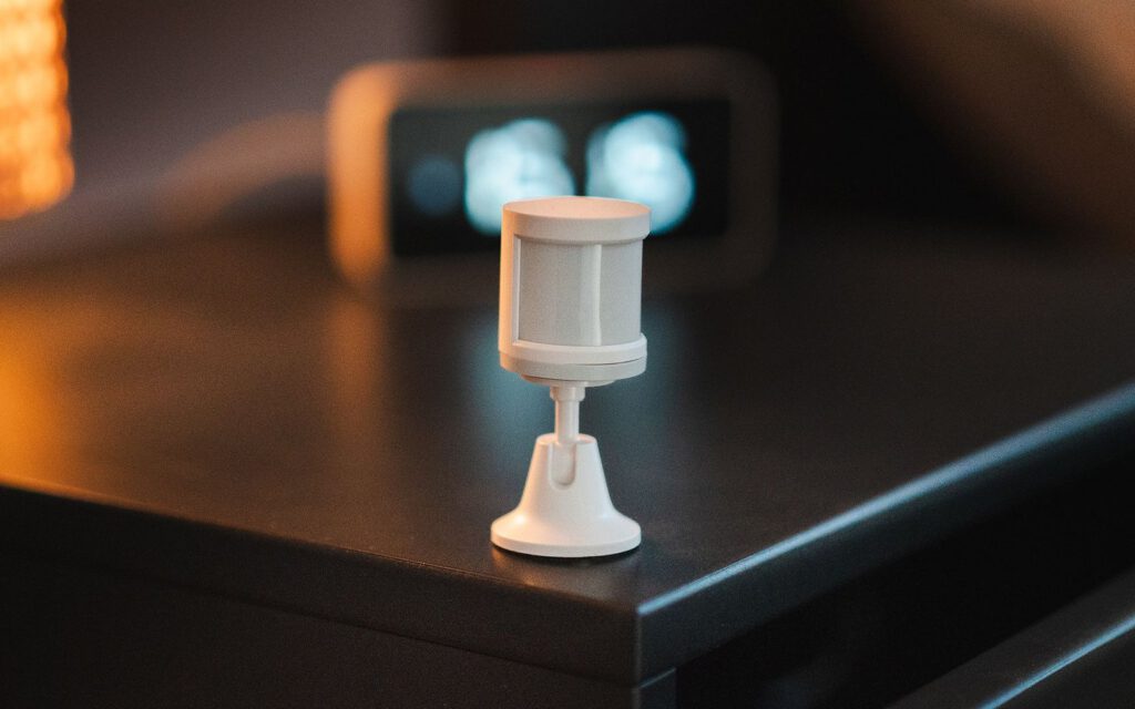 Witte bewegingssensor op een nachtkastje met lamp en klok op de achtergrond